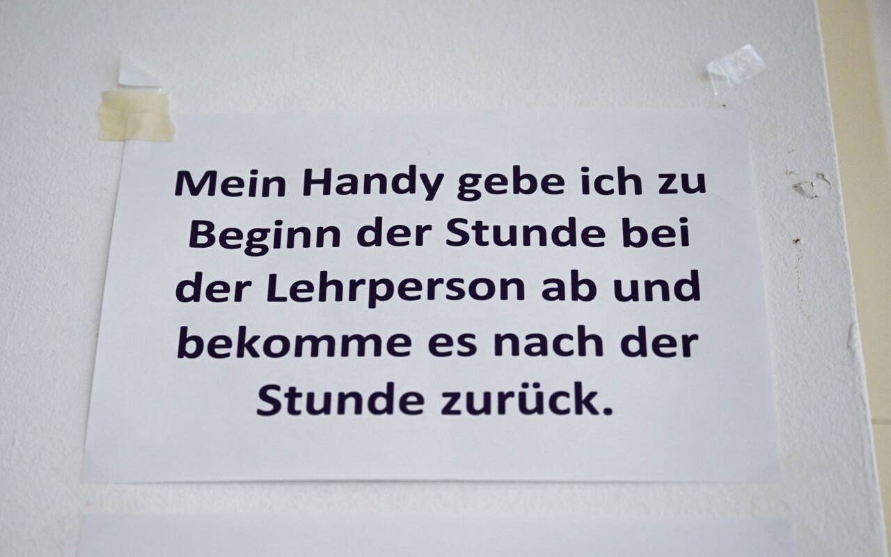 "Mein Handy gebe ich zu Beginn der Stunde bei der Lehrperson ab und bekomme es nach der Stunde zurück", steht in einem Klassenzimmer an einer Schule.